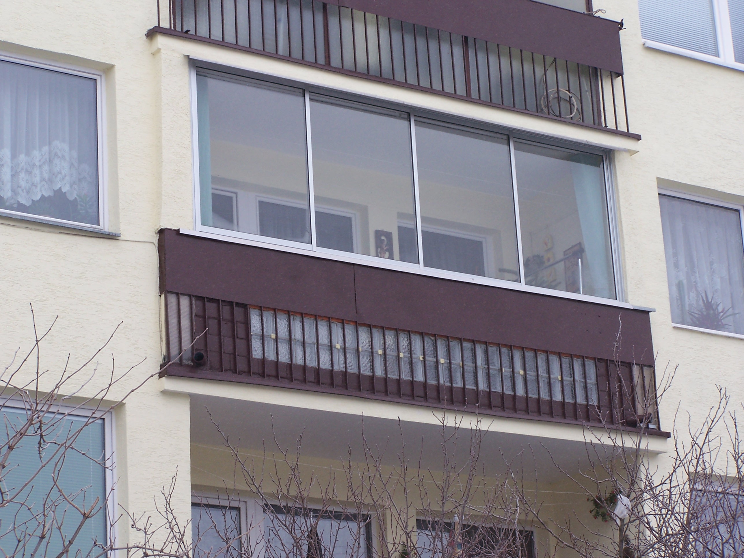 Zabudowany balkon z przesuwnymi oknami aluminiowymi, widok z dołu na kremową elewację budynku i metalową balustradę z dekoracyjnymi szybami.